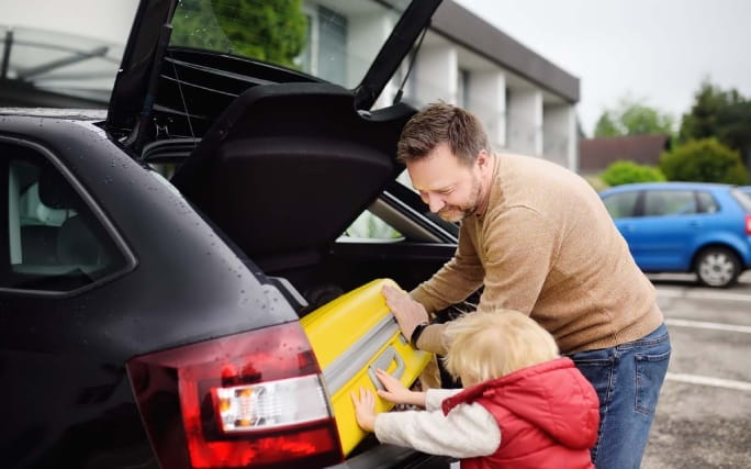 Pai põe uma bagagem na mala de um carro alugado ao lado do seu filho pequeno.