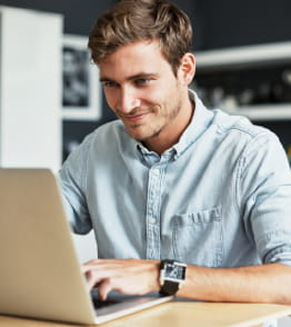 Homem jovem dentro de casa trabalha, sorridente, num portátil.
