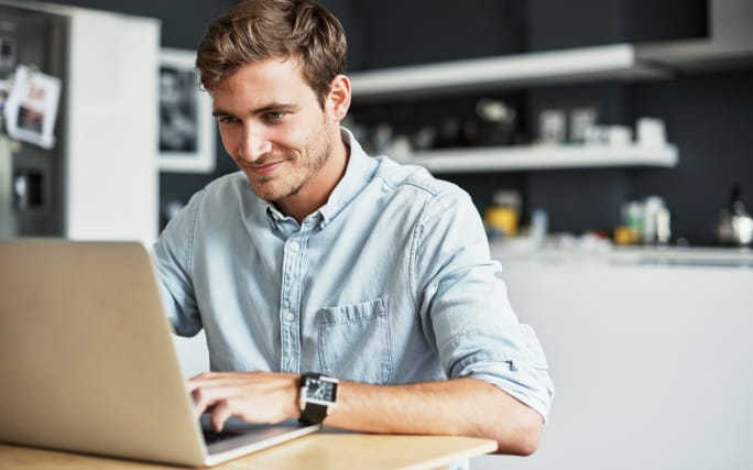 Homem jovem dentro de casa trabalha, sorridente, num portátil.