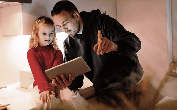 Pai e filha a observar conteúdo num tablet numa cozinha iluminada.