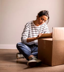 Jovem sentada no chão a escrever numa caixa de cartão durante uma mudança.