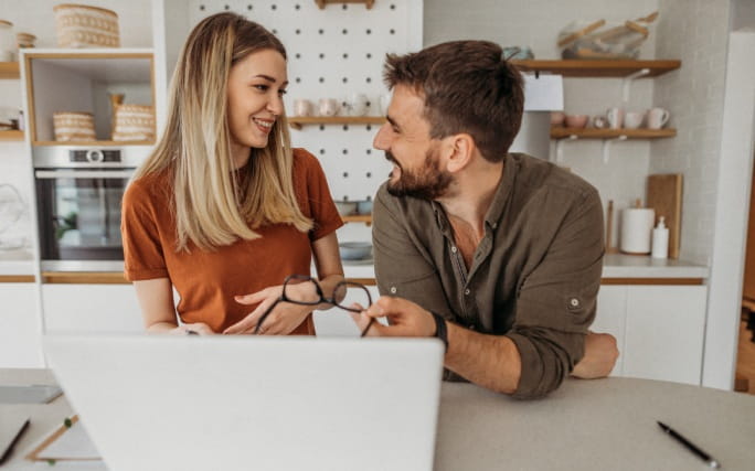 Um casal jovem numa cozinha, conversa animadamente de frente para um laptop aberto.