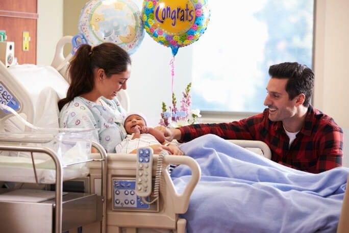 No quatro de um hospital, uma jovem mulher sentada na cama segura o primeiro filho ao colo e o pai faz-lhe uma festa.