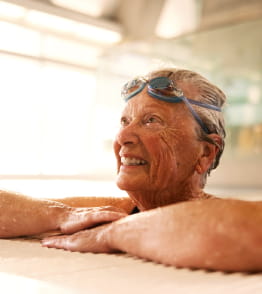 Mulher idosa a sorrir dentro de uma piscina interior com óculos de natação.