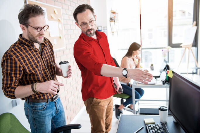 Dois homens no escritório a conversarem e a apontarem para um computador.